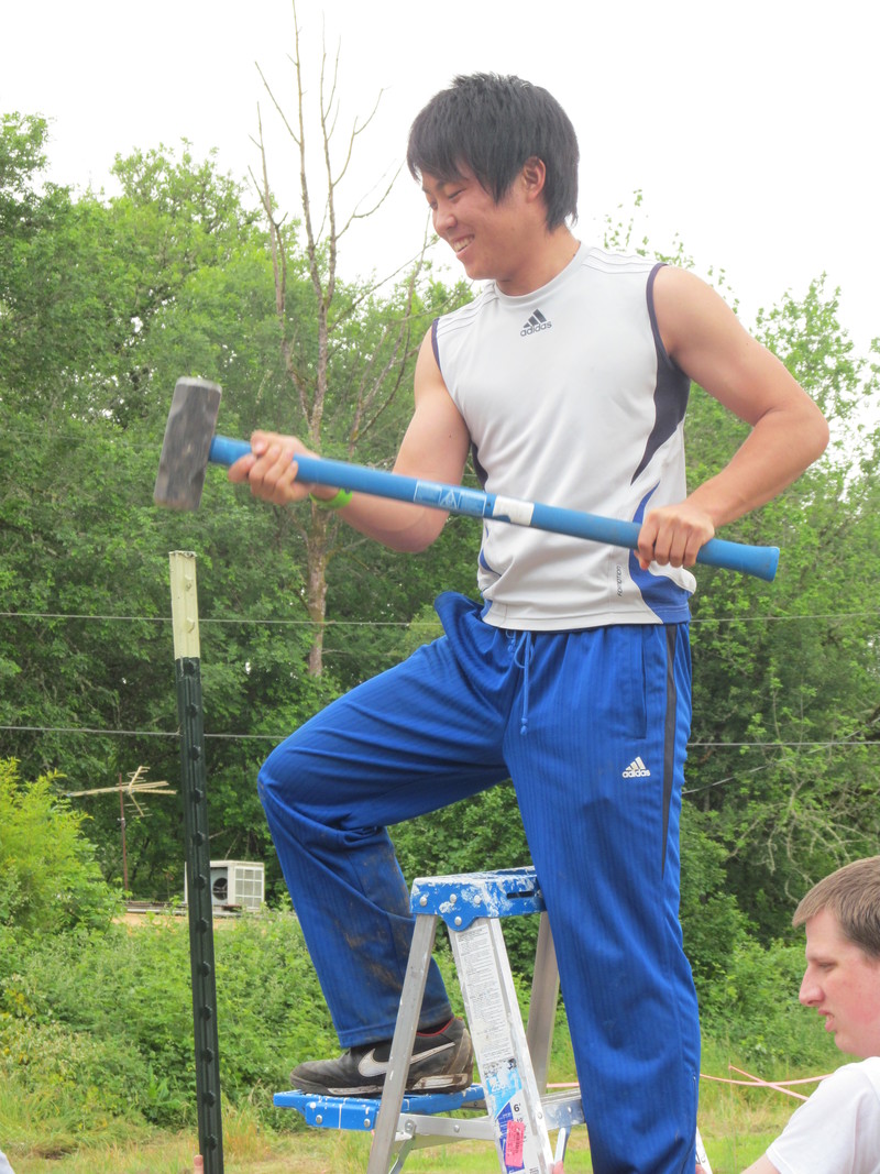 Kai pounds in a fence post. Isaac holds the ladder.